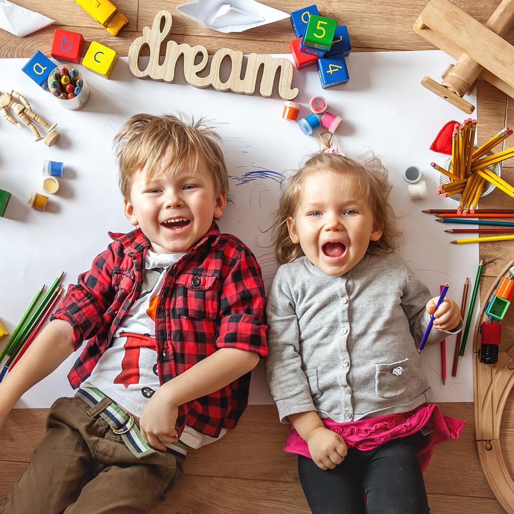 Zwei Kinder liegen fröhlich nebeneinander auf einem großen Blatt Papier, umgeben von bunten Bastelmaterialien wie Buntstiften, Holzbausteinen und Spielzeug. Über ihnen liegt das Wort „dream“ aus Holzbuchstaben.