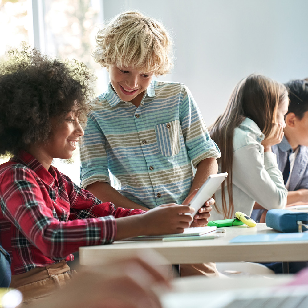 Eine Gruppe von Schülerinnen und Schülern in einem hellen Klassenzimmer. Zwei Kinder im Vordergrund, ein Junge mit lockigen, dunklen Haaren im karierten Hemd und ein blonder Junge im gestreiften Hemd, schauen auf ein Tablet und lachen. Im Hintergrund unterhalten sich andere Kinder an ihren Plätzen. Der Raum ist freundlich und modern gestaltet.