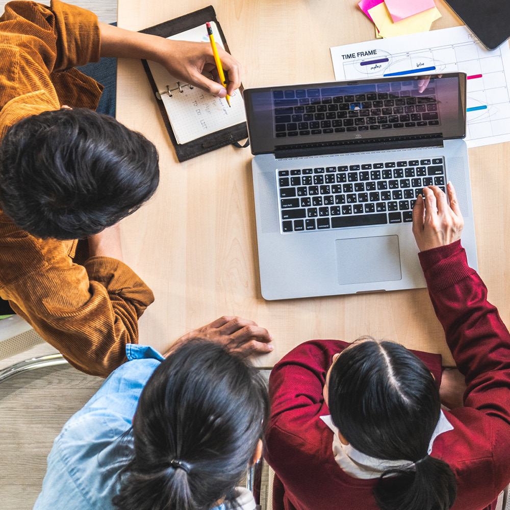 Drei Personen sitzen um einen Holztisch und arbeiten gemeinsam. In der Mitte steht ein Laptop, den alle ansehen. Einige machen Notizen in Notizbüchern, während andere Tablets oder Stifte benutzen. Auf dem Tisch liegen zusätzlich Dokumente und Schreibmaterialien. Die Ansicht ist von oben. Die Szene vermittelt eine produktive und kooperative Arbeitsatmosphäre.