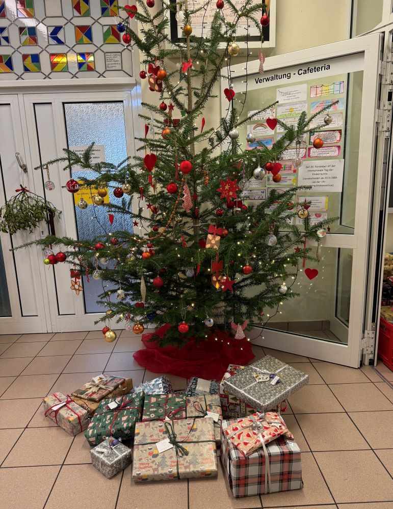 Ein geschmückter Weihnachtsbaum steht in einer Eingangshalle vor der Tür der Kolpingschule in Aschaffenburg. Die Tür hat die Beschriftung: "Verwaltung - Cafeteria". Der Baum ist mit roten und goldenen Kugeln, Herzschmuck, Lametta und Lichterketten dekoriert. Um den Baum herum liegen zahlreiche bunt verpackte Geschenkpäckchen in verschiedenen Größen und Mustern auf dem Boden. Im Hintergrund sind farbenfrohe Glasmosaiken zu sehen.