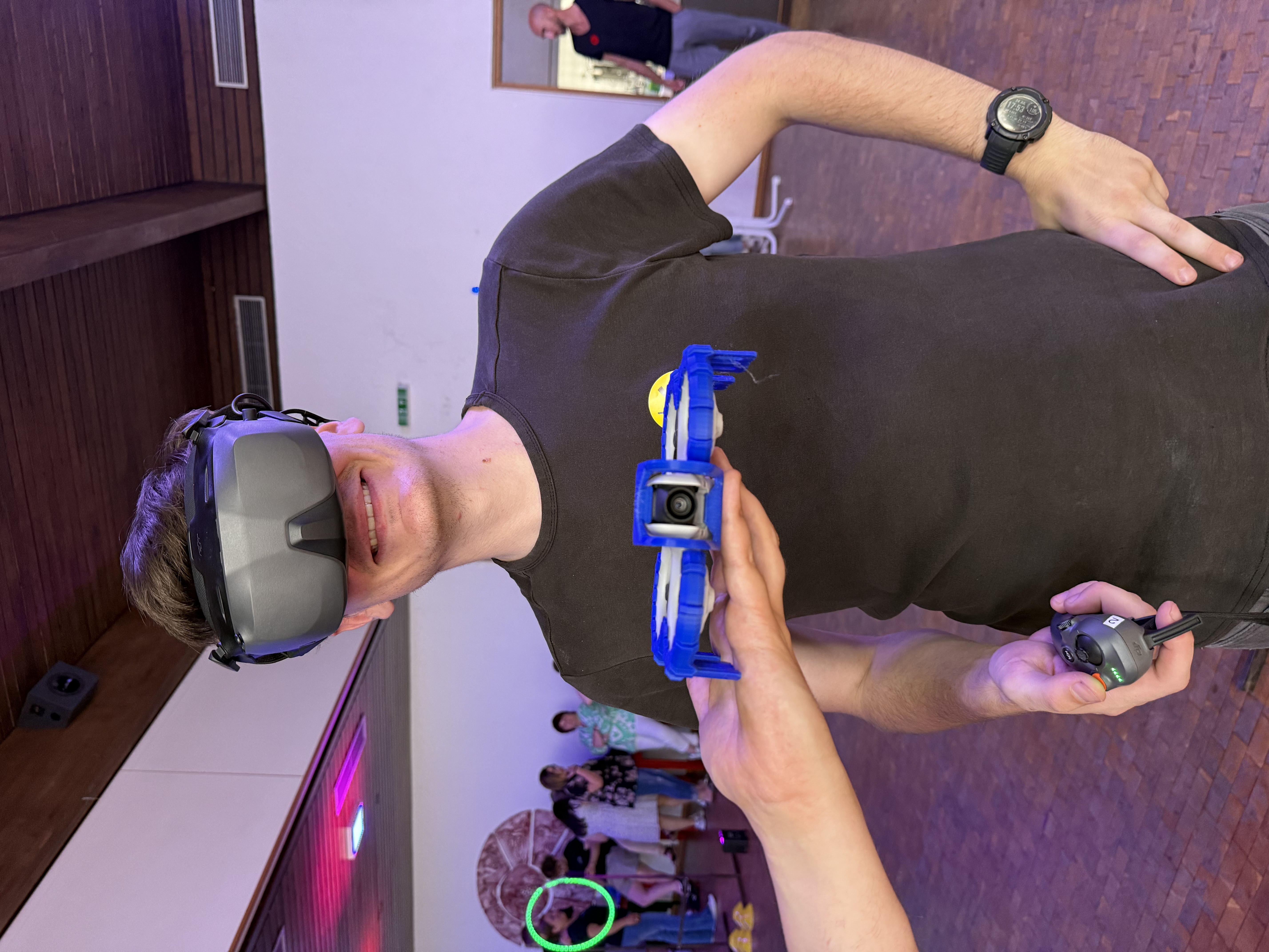 Ein junger Mann mit schwarzem T-Shirt trägt eine große Virtual-Reality-Brille und hält eine Fernbedienung in der Hand. Eine kleine blaue Drohne wird ihm von einer anderen Hand vor die Brust gehalten. Im Hintergrund sind Menschen in einem hellen Raum mit Holzboden zu sehen.