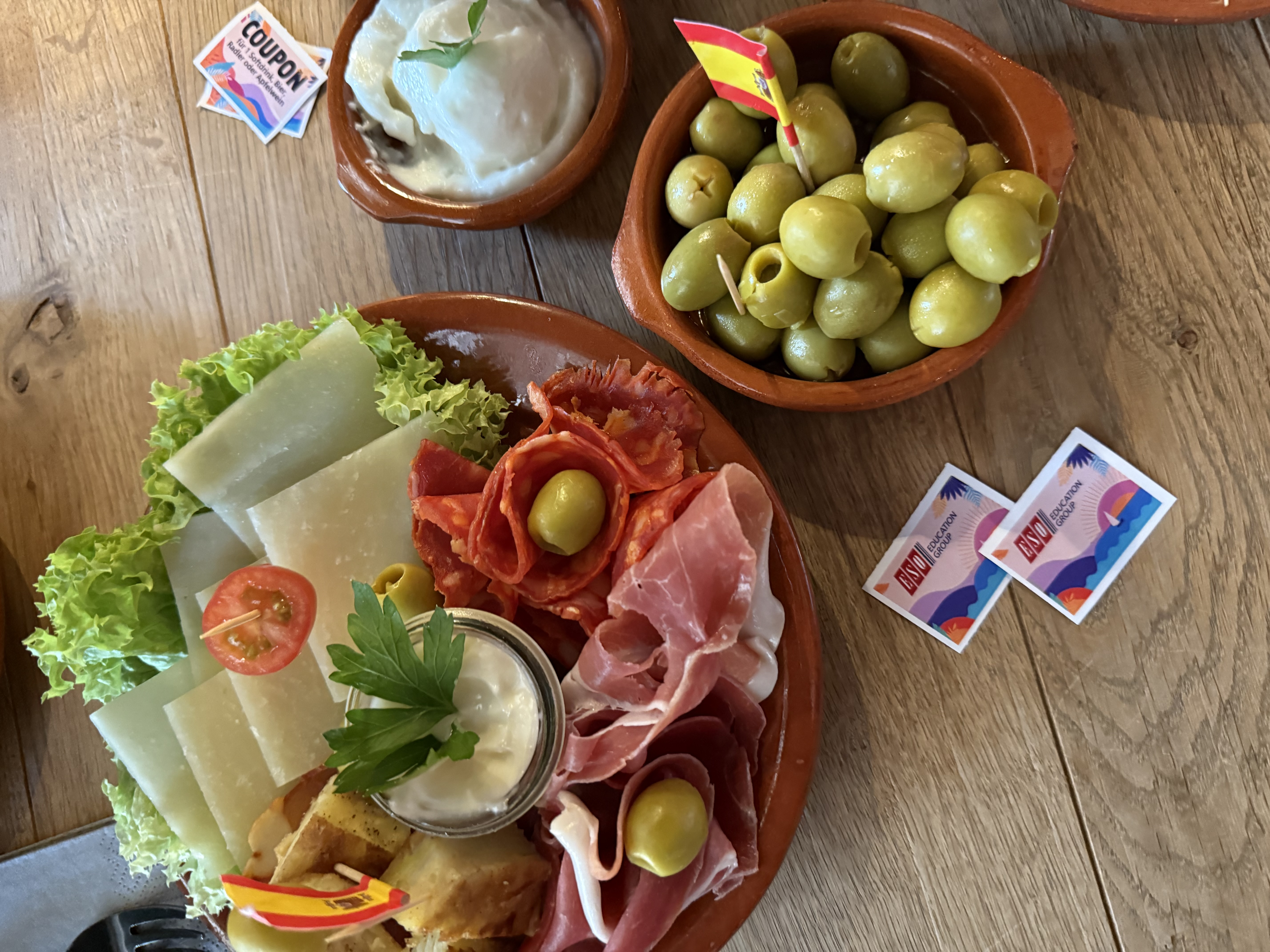 Ein rustikaler Holztisch mit einem spanischen Tapas-Teller: Käse, Schinken, Chorizo, Oliven, Brot und ein Dip sind appetitlich angerichtet. Daneben stehen kleine Schälchen mit grünen Oliven und Aioli. Auf dem Tisch liegen bunte Gutscheine mit der Aufschrift „ESO Education Group“.