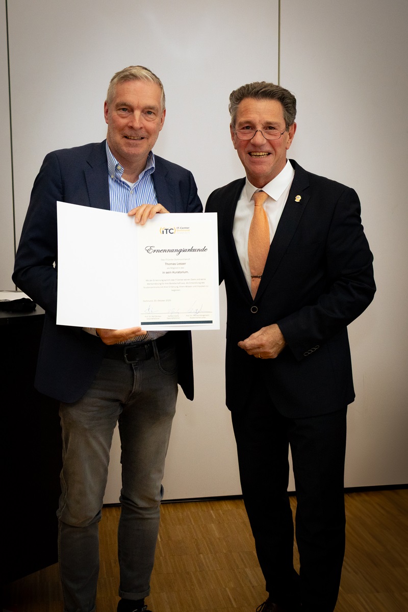 Zwei Männer stehen in einem hellen Veranstaltungsraum beim 25-jährigen Jubiläum des IT-Centers Dortmund. Prof. Dr. Ralf Brickau hält eine Ehrenurkunde der IHK Dortmund in den Händen, der andere steht daneben und lächelt. Beide tragen Anzug oder Businesskleidung. Die Urkunde ist gut sichtbar, aber die Personennamen sind nicht erkennbar wiedergegeben. Die Szene wirkt offiziell und würdevoll und zeigt die feierliche Übergabe einer Auszeichnung.