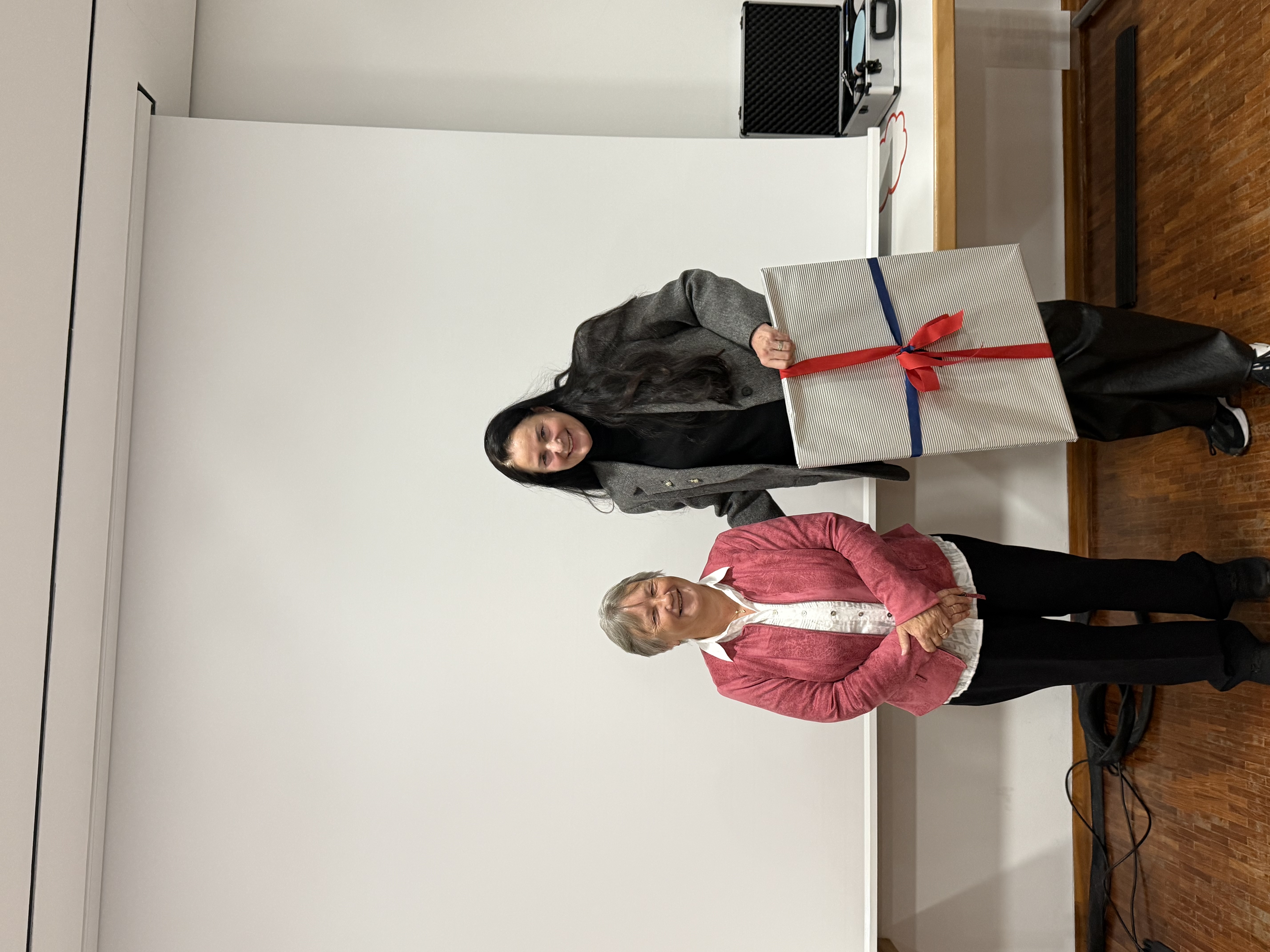 Zwei Frauen stehen in einem Seminarraum vor einer großen, weißen Projektionswand. Die kleinere Frau links trägt eine pinke Jacke über einer weißen Bluse und steht mit gefalteten Händen da. Die größere Frau rechts trägt einen grauen Blazer und schwarze Hose und hält ein großes, rechteckiges, eingepacktes Geschenk in der Hand.