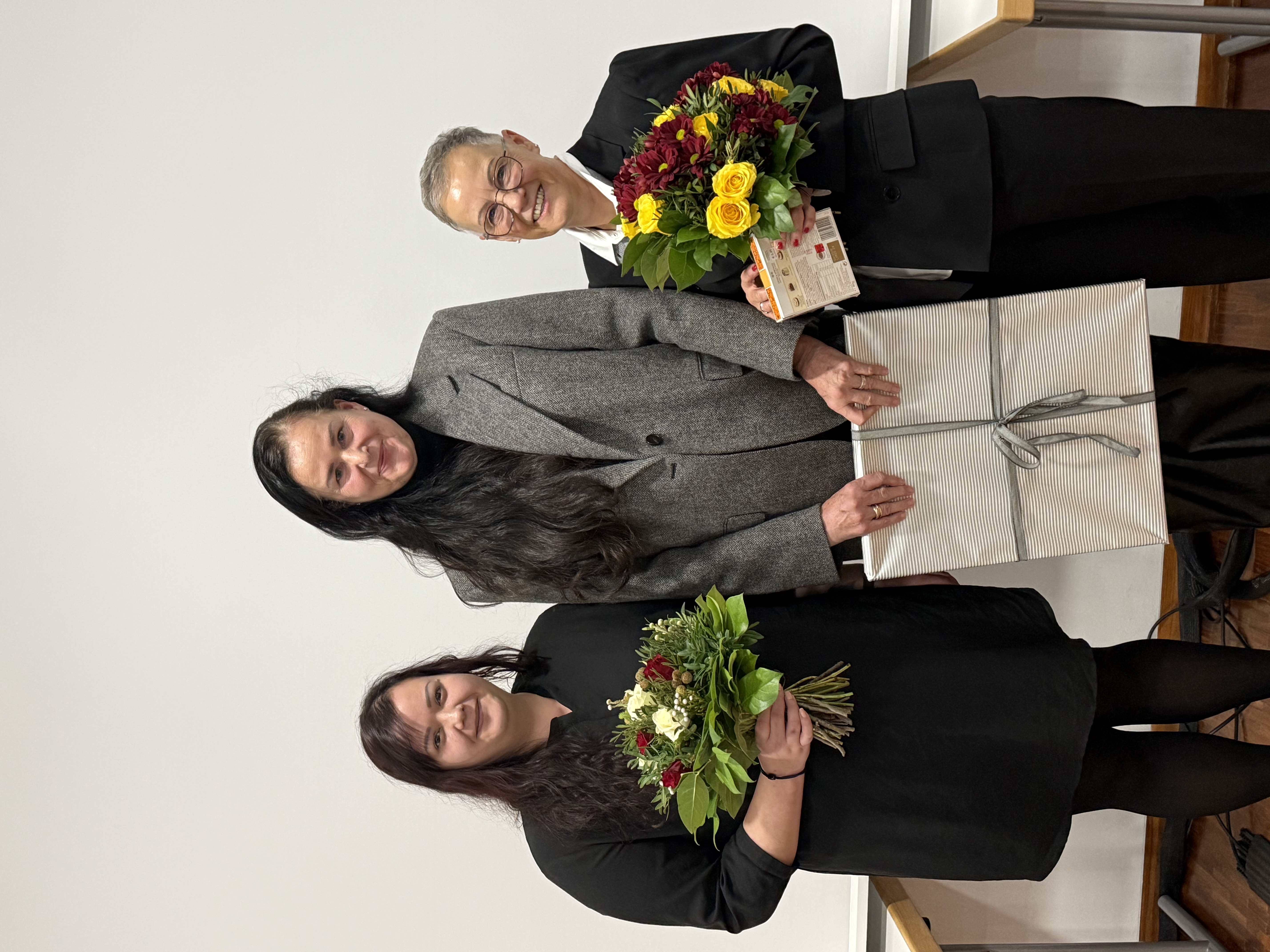 Drei Frauen stehen nebeneinander vor einer weißen Wand und schauen lächelnd in die Kamera; die Frau in der Mitte trägt einen grauen Blazer und hält ein großes, hell verpacktes Geschenk, während die Personen links und rechts in dunkler Kleidung jeweils einen farbigen Blumenstrauß in den Händen halten.