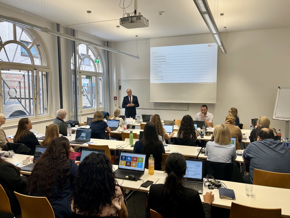 In einem Klassenzimmer steht Heinrich Geusken am Tisch des Dozenten vor den Teilnehmenden und spricht, während eine Präsentation an die Wand geworden wird. Die Teilnehmenden haben Laptops auf ihren Tischen stehen. Der Raum scheint ebenerdig zu sein, denn hinter den Fenstern sind Autos zu erkennen.