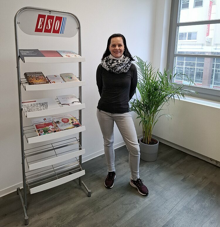 Mitarbeiter Nicole Höhnow posiert für ein Foto, im Hintergrund eine große Goldfruchtpalme in hellgrauem Übertopf und ein großes Regal mit Aufschrift ESO, in dem Informationsbroschüren ausliegen.
