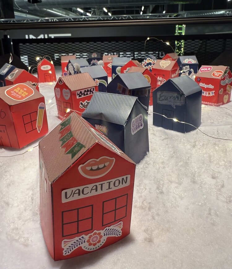 Kleine, bunt gestaltete Papierhäuser stehen dekorativ in künstlichem Schnee und werden von zarten Lichterketten beleuchtet. Die Häuschen tragen Aufkleber wie „Vacation“, „Good Vibes“ oder kleine Symbole. Die Szene entstand bei der Weihnachtsfeier 2025 der ESO Education Group bei der ZEEEM eSports GmbH und zeigt eine kreative, festliche DIY-Weihnachtslandschaft.