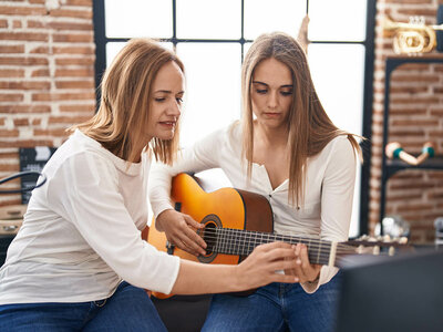 Zwei Frauen sitzen nebeneinander in einem Raum mit Ziegelsteinwänden und großen Fenstern. Die ältere Frau links zeigt der jüngeren Frau rechts, wie man Gitarre spielt. Beide tragen weiße Oberteile. Die ältere Frau hält die Gitarre und zeigt der jüngeren Frau, wie man die Saiten greift. Die jüngere Frau beobachtet konzentriert die Bewegungen. Im Hintergrund sind eine Trompete und andere Gegenstände zu sehen, was auf einen musikalischen oder kreativen Raum hindeutet. Beide wirken engagiert und geduldig.