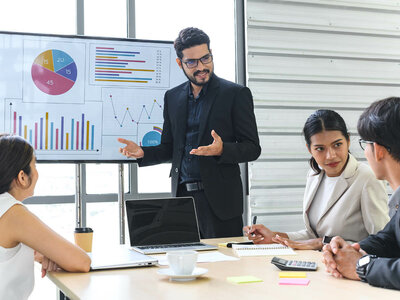 Ein Mann in einem Anzug präsentiert Daten auf einem Bildschirm mit Diagrammen und Grafiken während eines Meetings. Drei weitere Personen sitzen am Tisch, eine Frau und zwei Männer, die aufmerksam zuhören und Notizen machen. Auf dem Tisch stehen ein Laptop, ein Kaffeebecher und ein Taschenrechner.