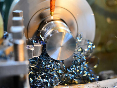 Detailaufnahme eines Metallbearbeitungsprozesses an einer Drehmaschine. Ein zylindrisches Metallstück rotiert mit hoher Geschwindigkeit, während ein Schneidwerkzeug es formt. Um das Werkstück herum sind Metallspäne zu sehen, was auf eine aktive Bearbeitung hinweist.