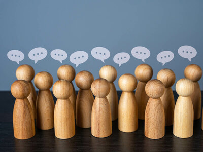 Eine Gruppe von hölzernen Figuren steht in einer Reihe auf einer dunklen Oberfläche. Über jeder Figur schwebt eine weiße Sprechblase mit drei Punkten, die eine Diskussion oder einen Dialog symbolisieren. Der Hintergrund ist in einem blauen Farbton gehalten.