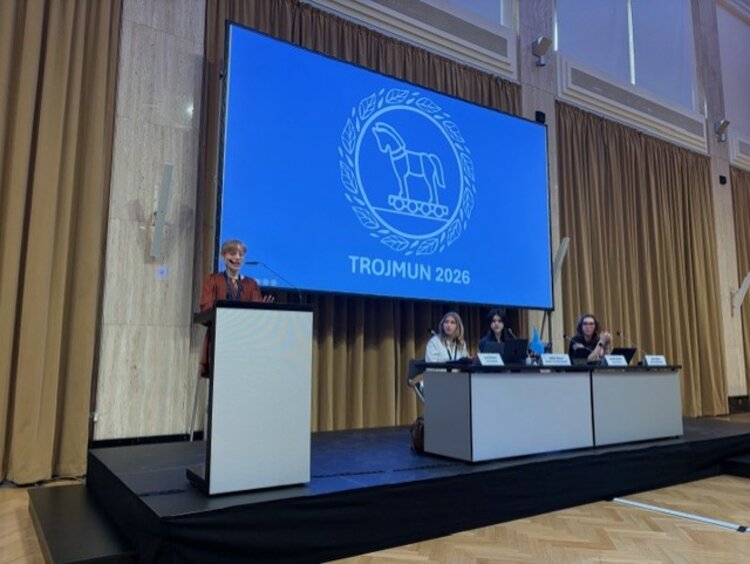 Eine Person hält eine Rede an einem Rednerpult auf einer Bühne. Im Hintergrund zeigt eine große Leinwand das Logo und den Schriftzug „TROJMUN 2026“. Drei Personen sitzen an einem Tisch rechts auf der Bühne.