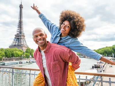 Zwei Personen stehen vor dem Eiffelturm in Paris. Eine hebt die Arme. Die Seine fließt unter ihnen. Helle Wolken am Himmel.