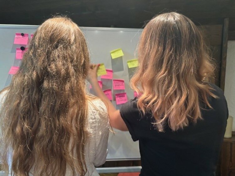Zwei Frauen stehen mit dem Rücken zur Kamera vor einem Whiteboard und heften pinke und gelbe Post-its an. Ihre konzentrierte Haltung zeigt den kollaborativen Charakter der Teamarbeit während des Workshops.