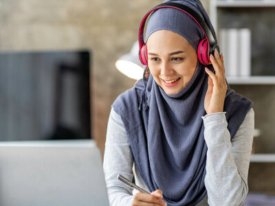 Eine lächelnde Frau mit Kopfhörern und einem grauen Hijab sitzt an einem Schreibtisch und hält einen Stift in der Hand. Im Hintergrund sind ein Laptop und Bücherregale zu sehen.