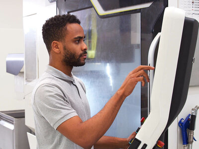 Ein Mann mit einem grauen Polohemd steht vor einer großen industriellen Maschine und bedient das Bedienfeld. Er scheint konzentriert und drückt einen Knopf oder Schalter an der Maschine. Die Maschine hat eine große Tür und verschiedene Bedienelemente. Im Hintergrund sind weitere Maschinen und Geräte zu sehen, die auf eine industrielle Umgebung hinweisen.