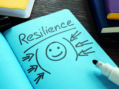 Ein Notizbuch mit einer blauen Seite, auf der das Wort 'Resilience' (Widerstandsfähigkeit) geschrieben steht. Unter dem Wort ist ein lächelndes Gesicht gezeichnet, umgeben von Pfeilen, die auf das Gesicht zeigen. Ein weißer Marker mit schwarzer Spitze liegt auf der Seite des Notizbuchs. Im Hintergrund sind verschiedene farbige Notizbücher und Stifte zu sehen.