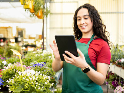 Eine junge Frau mit lockigem Haar und einer grünen Schürze über einem roten Shirt steht in einem Blumenladen. Sie hält ein Tablet in den Händen und lächelt. Um sie herum sind verschiedene Blumen und Pflanzen in Töpfen zu sehen. Der Hintergrund zeigt Regale mit weiteren Blumen und ein helles Gewächshaus.