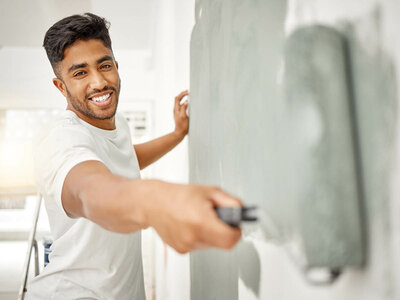 Eine Person mit streicht eine Wand mit einer Farbrolle. Die Person trägt ein weißes T-Shirt und scheint gerade graue Farbe auf die Wand aufzutragen.