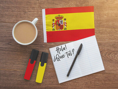 Ein Foto von einem Holztisch mit einer Tasse Kaffee, einer kleinen spanischen Flagge, zwei Textmarkern in den Farben der Flagge und einem Notizblock, auf dem 'Hola! ¿Qué tal?' geschrieben steht.