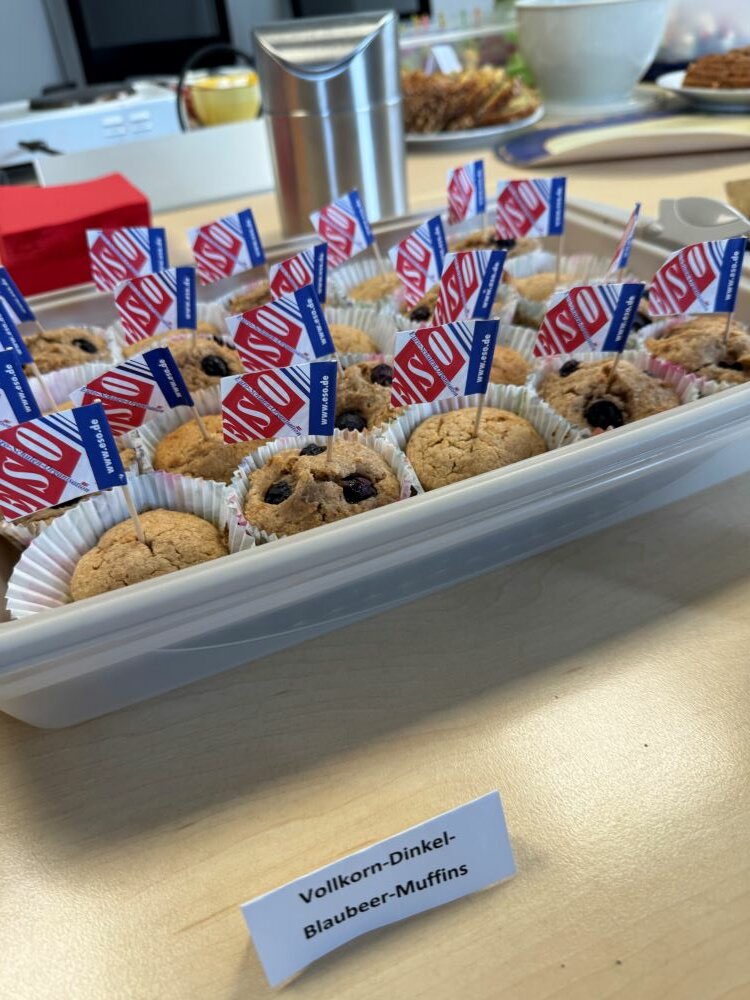 Achtsamtkeit im Fokus: ESO Education Group veranstaltet erneut erfolgreichen Gesundheitstag in Stockstadt Muffins in einer offenen weißen Box, die mit kleinen Fähnchen verziert sind. Daneben liegt ein Schild mit der Aufschrift „Vollkorn-Dinkel-Blaubeer-Muffins“.