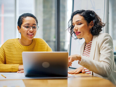 Zwei lächelnde Frauen sitzen an einem Tisch und arbeiten gemeinsam an einem Laptop. Die Frau links trägt eine gelbe Strickjacke und eine Brille, während die Frau rechts ein weißes Blazer-Jacket über einem rot-weiß gestreiften Shirt trägt. Beide wirken engagiert und gut gelaunt. Der Hintergrund zeigt große Fenster, die viel Tageslicht in den Raum lassen, möglicherweise ein Büro oder ein Besprechungsraum.