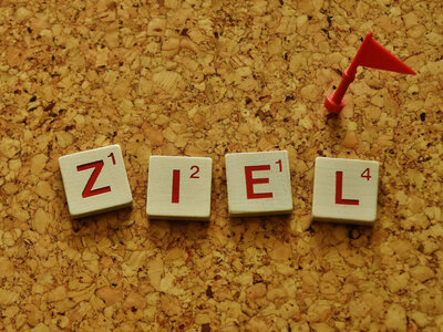 Vier Scrabble-Steine auf einer Korkplatte. Die Steine zeigen die Buchstaben Z, I, E und L mit verschiedenen Zahlenwerten: Z^1, I^2, E^1 und L^4. Ein roter Spielstein mit einer kleinen Flagge ist ebenfalls auf der Platte zu sehen.