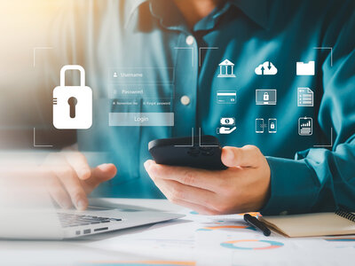 Eine Person sitzt an einem Laptop und hält ein Smartphone in der Hand. Über dem Smartphone schwebt ein holografisches Login-Fenster mit Feldern für Benutzername und Passwort sowie einem Login-Button. Das Symbol eines Vorhängeschlosses ist über dem Login-Fenster zu sehen, was auf Sicherheit hinweist.