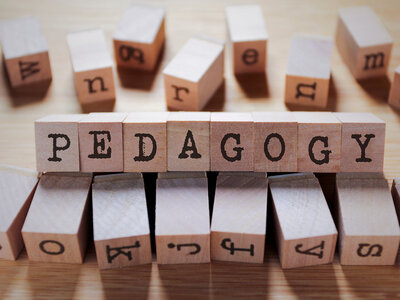 Holzblöcke mit Buchstaben, die das Wort 'PEDAGOGY' bilden. Die Blöcke sind auf einer Holzoberfläche angeordnet, und im Hintergrund sind weitere Holzblöcke mit verschiedenen Buchstaben zu sehen.