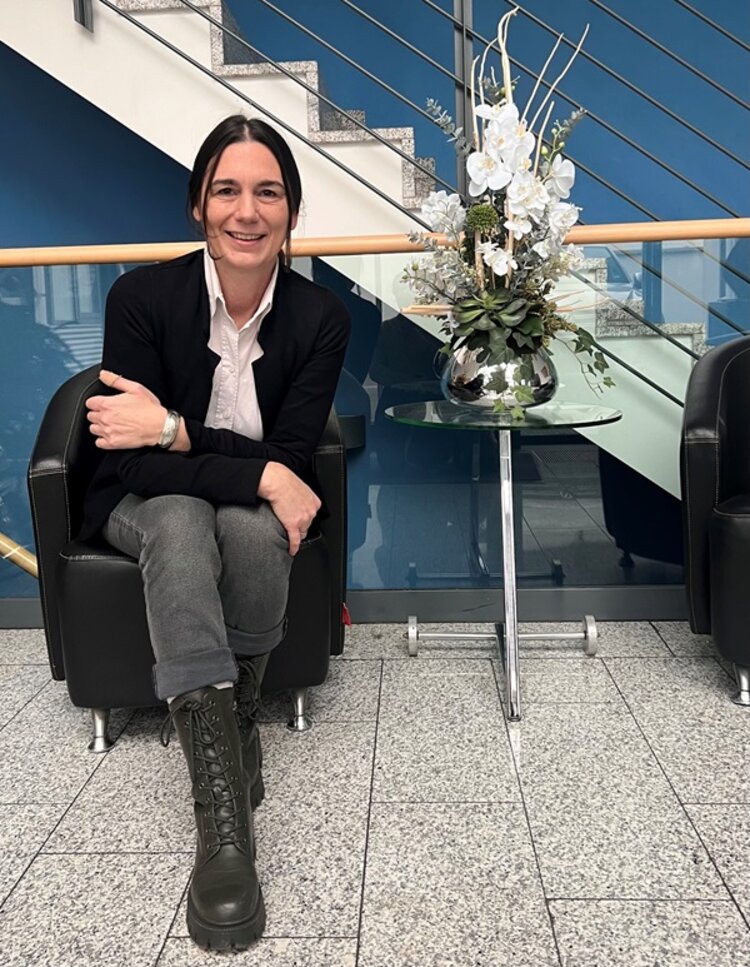 Mitarbeiterin Katrin Menzer sitzt auf schwarzem Sessel im Treppenhaus, neben ihr ein Glastisch mit Vase und weißen Schnittblumen.