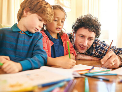 Ein Vater mit lockigen Haaren und einem karierten Hemd sitzt mit seinen beiden Kindern an einem Tisch und macht Hausaufgaben oder kreative Arbeiten. Der Junge links trägt ein blaues Langarmshirt mit Streifen und hält einen Stift in der Hand. Das Mädchen in der Mitte trägt ein dunkles Oberteil mit einer roten Jacke und malt konzentriert. Auf dem Tisch liegen viele bunte Stifte und Blätter. Alle drei wirken engagiert und konzentriert. Der Raum ist hell und freundlich.