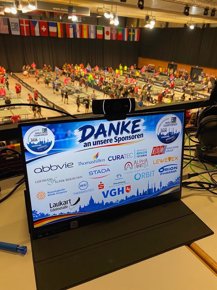 Gemeinsam im Einsatz für die PingPongParkinson-WM 2026 Auf einem Bildschirm sind unter der Überschrift "Danke an unsere Sponsoren" verschiedene Unternehmenslogos aufgelistet. Hinter dem Bildschirm ist eine Sporthalle mit PingPong-Spielern und internationalen Flaggen zu sehen.