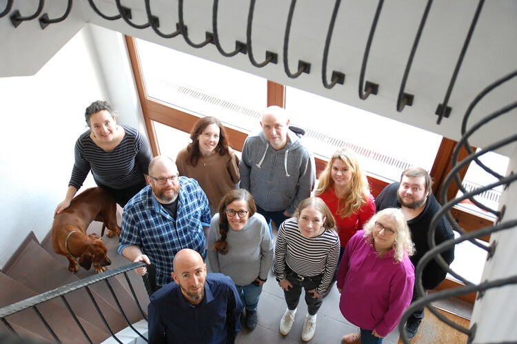Das Foto zeigt das gesamte ECC-Team im Treppenhaus des Firmengebäudes. Die Gruppe steht im Erdgeschoss, das Bild ist aus dem ersten Stock nach unten aufgenommen. Es zeigt sechs Mitarbeiterinnen und vier Mitarbeiter sowie einen braunen Hund der Rasse Rhodesian Ridgeback.