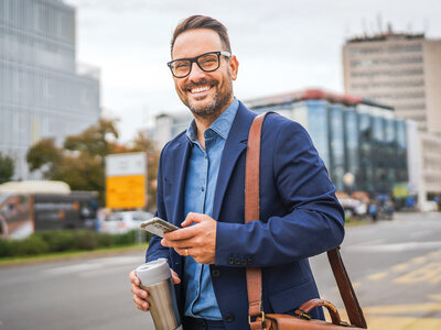 Ein Mann läuft an einer Straße in einer belebten städtischen Umgebung entlang. Er trägt eine dunkle Brille, einen blauen Anzug mit einem hellblauen Hemd und hat eine braune Ledertasche über der Schulter. In der linken Hand hält er eine wiederverwendbare Kaffeetasse, in der rechten Hand ein Smartphone, auf das er blickt. Im Hintergrund sind moderne Gebäude, ein Bus und weitere Elemente einer wachen Stadtlandschaft zu erkennen. Der Mann wirkt entspannt und selbstbewusst, während er sich auf sein Smartphone konzentriert. Die morgendliche Atmosphäre ist urban und dynamisch.