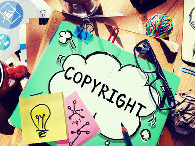 Ein grüner Ordner mit dem Wort 'COPYRIGHT' in einer Sprechblase liegt auf einem Schreibtisch. Auf dem Ordner befinden sich eine Brille, Büroklammern und ein gelber Notizzettel mit einer Glühbirne darauf. Um den Ordner herum liegen weitere Notizzettel, ein roter Tacker, ein Knäuel bunter Gummibänder und andere Büromaterialien.