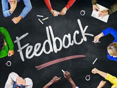 Eine Gruppe von Personen sitzt um eine Tafel herum, auf der das Wort 'Feedback' in großen, weißen Buchstaben geschrieben steht. Die Personen sind in verschiedenen Posen und scheinen sich auf die Tafel zu konzentrieren. Einige halten Stifte oder Kreide, was darauf hindeutet, dass sie an einer Feedback-Session oder einem Brainstorming teilnehmen. Auf der Tafel liegen auch verschiedene farbige Kreidestücke.
