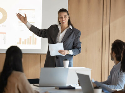 In einem modernen Besprechungsraum hält eine Frau in einem dunklen Blazer und mit einem weißen Blatt Papier in der Hand eine Präsentation. Sie steht vor einer Leinwand, auf der ein Diagramm mit einem Kreis- und einem Balkendiagramm zu sehen ist. Die Frau wirkt selbstbewusst und erklärt etwas, während sie mit der freien Hand eine Geste macht. Vor ihr sitzen drei Personen, die aufmerksam zuhören und Notizen auf ihren Laptops machen. Auf dem Tisch stehen Kaffeebecher. Der Raum ist mit hellen Holzwänden ausgestattet und wirkt professionell.