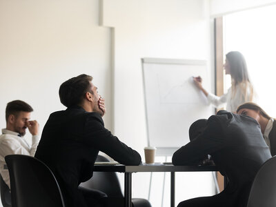 Ein Besprechungsraum, in dem mehrere Personen an einem Tisch sitzen. Einige der Personen sehen müde oder gelangweilt aus, während eine Frau im Hintergrund an einem Flipchart steht und etwas präsentiert. Auf dem Tisch stehen ein Kaffeebecher und einige Papiere.
