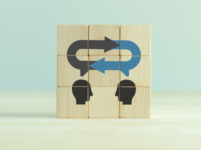 Ein Holzpuzzle, das aus mehreren Teilen besteht und eine Kommunikation zwischen zwei Personen darstellt. Das Puzzle zeigt zwei Köpfe, die sich gegenüberstehen, mit Sprechblasen, die durch Pfeile verbunden sind. Ein schwarzer Pfeil zeigt nach rechts und ein blauer Pfeil zeigt nach links, was auf einen Dialog oder eine Interaktion zwischen den beiden Personen hinweist.