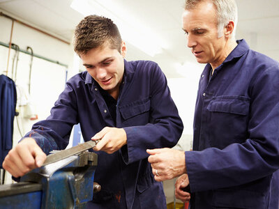 Zwei Männer in blauen Arbeitsanzügen stehen in einer Werkstatt. Der jüngere Mann hält ein Metallstück und arbeitet an einem Schraubstock. Der ältere Mann beobachtet und gibt möglicherweise Anweisungen oder Unterstützung. Im Hintergrund sind Werkzeuge und Geräte zu sehen, die auf eine industrielle oder handwerkliche Umgebung hinweisen.