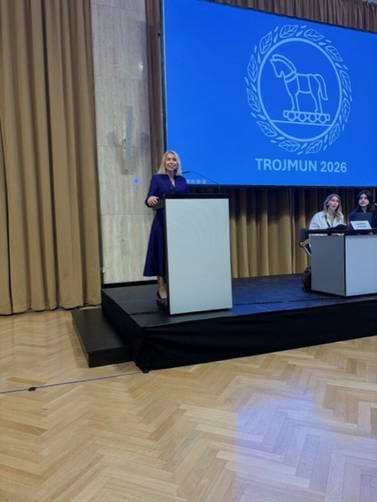 Eine Frau in einem dunkelblauen Kleid steht an einem Rednerpult auf einer Bühne. Im Hintergrund ist eine große Leinwand mit dem Logo und dem Schriftzug „TROJMUN 2026“ zu sehen. Zwei weitere Personen sitzen an einem Tisch rechts auf der Bühne.