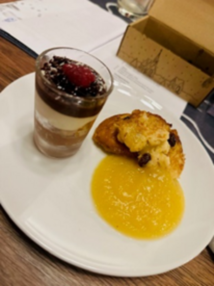 Weißer Dessertteller mit einem geschichteten Schokoladencreme-Glas mit Himbeere und einem Stück süßem Auflauf oder Hefezopf, serviert mit gelber Fruchtsauce.