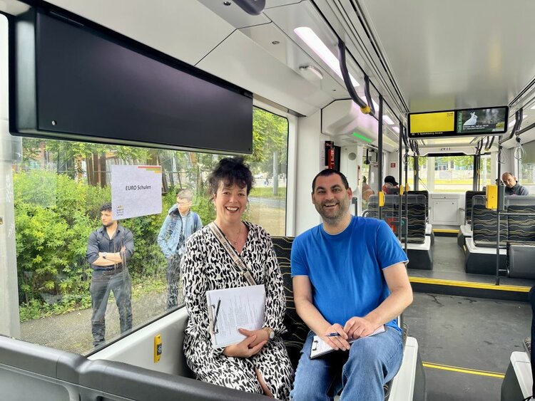 Zwei Mitarbeitende der ESO Education Group sitzen mit Unterlagen in einer modernen Straßenbahn und lächeln in die Kamera. Hinter dem Fenster ist ein Schild mit der Aufschrift „EURO Schulen“ sowie wartende junge Menschen zu sehen.