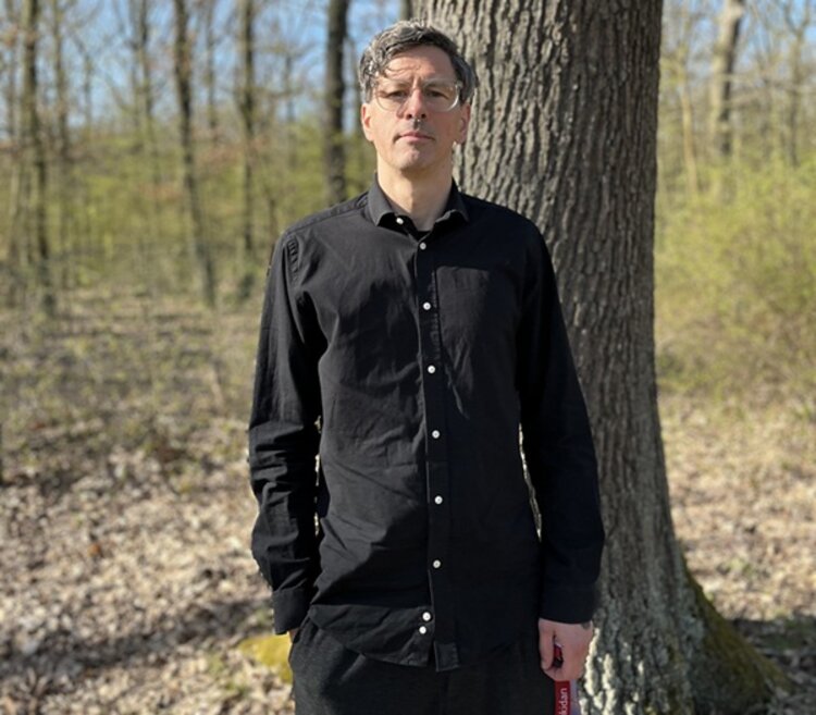 KITA Waldräuber Mitarbeiter posiert im anliegenden Wald für ein Foto, er trägt ein dunkles Hemd, eine dunkle Hose und eine Brille.