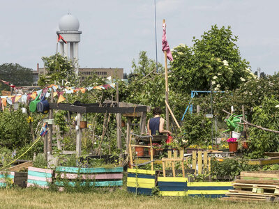Gemeinschaftsgarten auf dem Tempelhofer Feld in Berlin mit bunt bemalten Hochbeeten, Wimpelketten und verschiedenen Pflanzen. Eine Person arbeitet im Garten, während im Hintergrund der ehemalige Flughafen Tempelhof mit seiner Radarkuppel zu sehen ist.