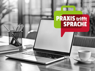 Auf dem Bild ist ein Schreibtisch in einem Büro zu sehen. Auf dem Tisch steht ein offener Laptop, daneben liegen ein Notizbuch und ein Behälter mit Stiften. Rechts vom Laptop steht eine Tasse auf einem Unterteller. Über dem Bild schwebt eine grüne Sprechblase mit dem Text "PRAXIS trifft SPRACHE" in weißer und roter Schrift. Der Hintergrund ist leicht unscharf, zeigt aber ein Büroumfeld.