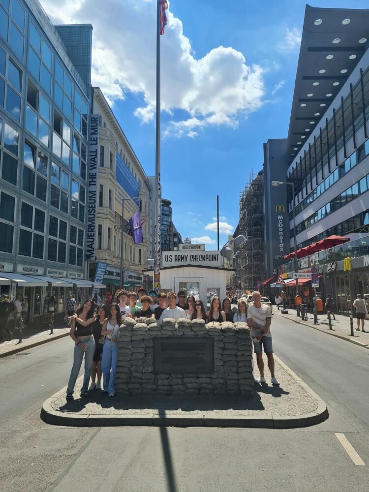 Klassenfahrt der Euroskola Prag nach Berlin Eine Gruppe von Jugendlichen und Erwachsenen steht vor dem Checkpoint Charlie in Berlin. Im Hintergrund sind moderne Gebäude, eine McDonald’s-Filiale und ein Banner mit der Aufschrift „The Wall Museum“ zu erkennen. Die Gruppe posiert vor einer Nachbildung der Sandacksäcke, die den historischen Kontrollpunkt markieren.