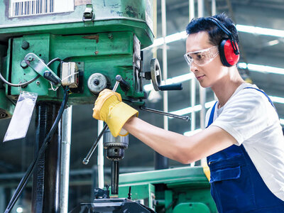 Eine Person bedient eine grüne industrielle Bohrmaschine in einer Werkstatt- oder Fabrikumgebung. Sie trägt gelbe Handschuhe, roten Gehörschutz und einen blauen Overall über einem weißen Hemd. Das Gesicht der Person ist aus Datenschutzgründen unscharf.