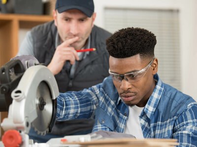 Zwei Personen befinden sich in einer Werkstatt. Eine Person bedient eine Kreissäge, während die andere Person zuschaut und einen roten Stift hält. Die Gesichter beider Personen sind aus Datenschutzgründen unscharf.